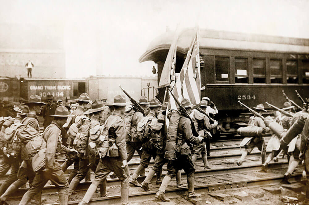 A 1917 New York National Guard regiment walks along railroad tracks toward railroad passenger cars on their way to training camp.